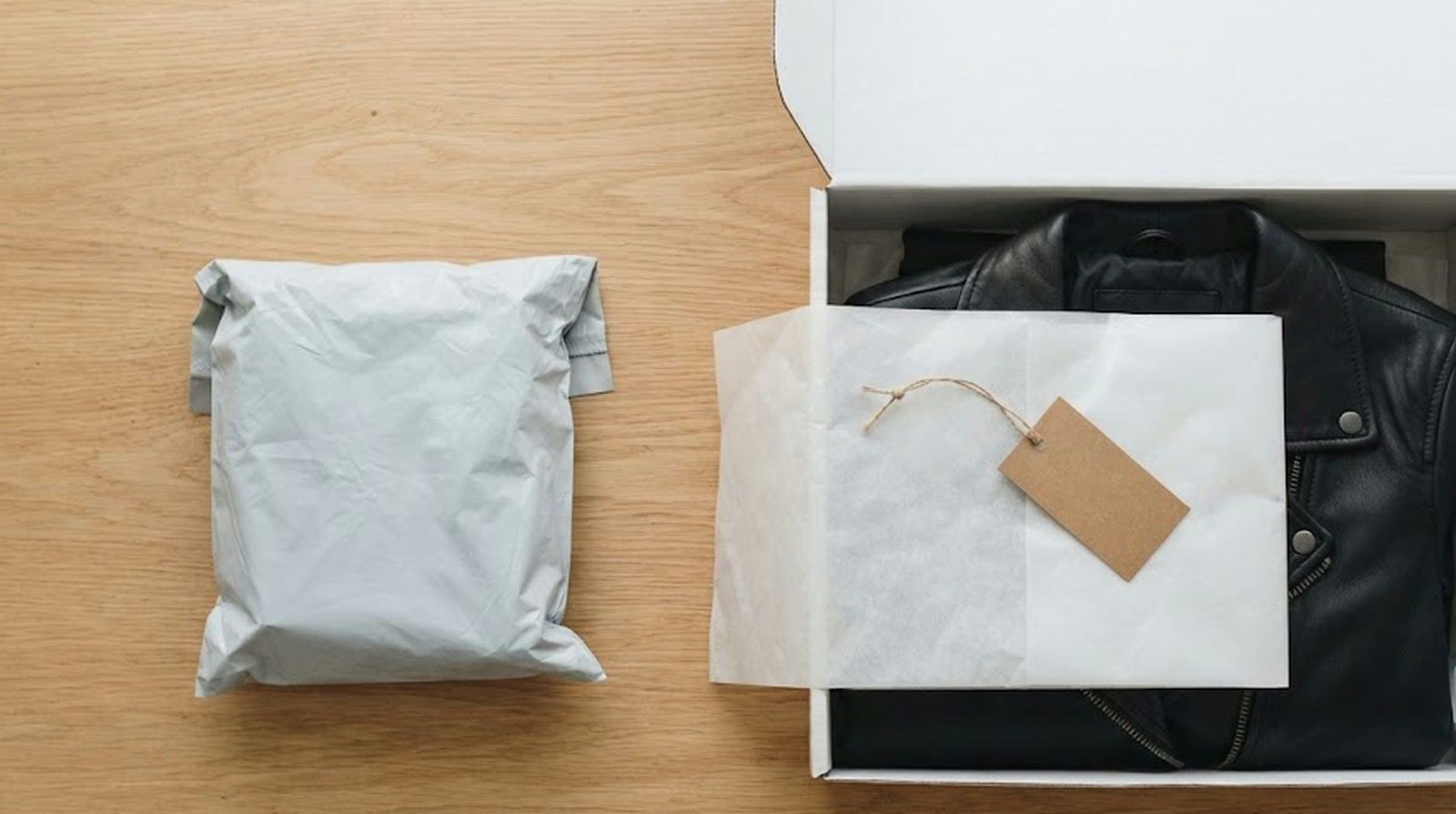 A plain grey polybag parcel beside an open white shipping box holding a folded leather jacket in tissue paper with a blank hang tag